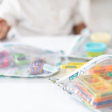 Close-up of a person with Bumkins Clear Travel Bag 3-Pack: Cacti filled with colorful toys and playdough on a white table.