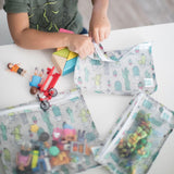 A child plays with toys and Bumkins Clear Travel Bag 3-Pack: Cacti on a white table.