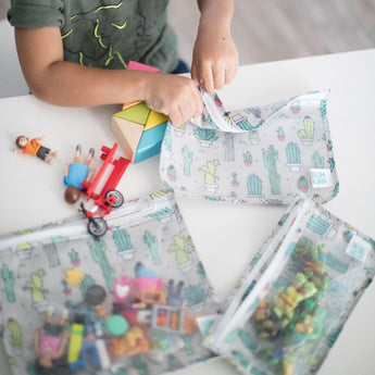 A child plays with toys and Bumkins Clear Travel Bag 3-Pack: Cacti on a white table.