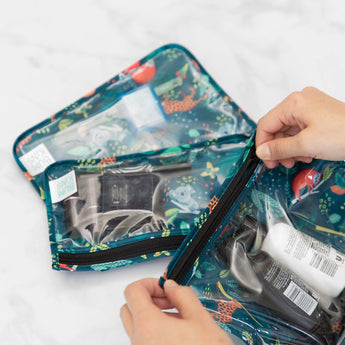 Hands opening a Bumkins Clear Travel Bag from the Jungle 3-Pack, showcasing toiletries on a soft white surface.