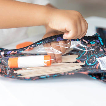 A child explores a Bumkins Clear Travel Bag from the Channel Kindness 3-Pack, discovering glue and craft sticks on a white surface.