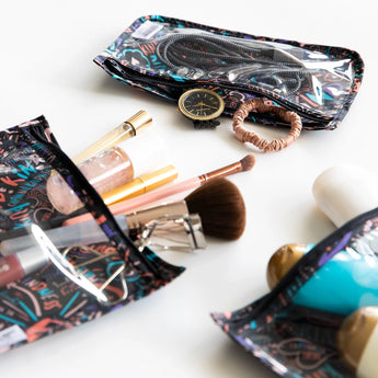 On a white surface, three TSA compliant pouches from the Bumkins Clear Travel Bag 3-Pack Channel Kindness display colorful patterns. One holds makeup and brushes, another carries a watch and hair accessories, while the third stores miscellaneous toiletries.