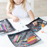 A child in a white shirt is immersed in drawing with colorful markers at a table. Nearby, three Bumkins Clear Travel Bags from the Channel Kindness 3-Pack are filled with art supplies.