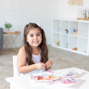 A young girl with long hair smiles at a table with colorful papers and crayons, her Bumkins Clear Travel Bag: Princess Magic nearby.