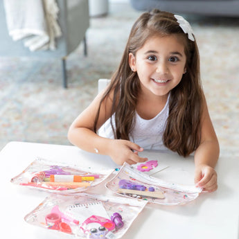 Young girl smiles at a table with Bumkins Clear Travel Bag 3 Pack: Princess Magic, featuring TSA-compliant craft supplies.