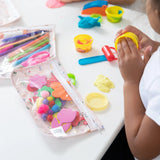 A child enjoys modeling clay on a white table, with creations stored in Bumkins Princess Magic Clear Travel Bag 3 Pack for future play.