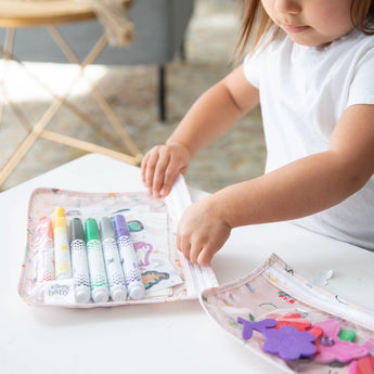 A child organizes markers and stickers in a Bumkins Clear Travel Bag from the Princess Magic pack on a white table.