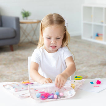 Young girl crafting happily, surrounded by Bumkins lightweight, waterproof Clear Travel Bag 3 Pack: Princess Magic.