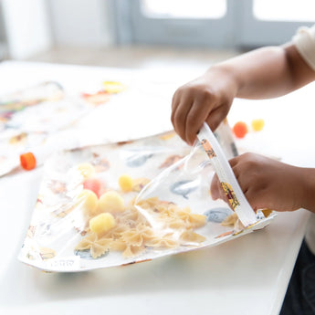 A child plays with pasta and pom-poms for sensory fun using Bumkins Clear Travel Bag 3-Pack: Winnie and Friends.