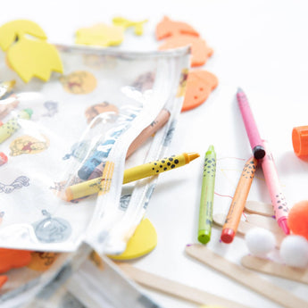 Colorful pencils and crayons near a Bumkins Clear Travel Bag with Winnie and Friends stickers.