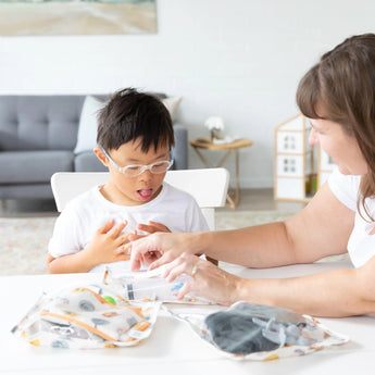 A child with glasses and an adult explore education materials in a cozy living room using Bumkins Clear Travel Bag 3-Pack: Winnie and Friends.