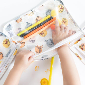 A childs hands arrange colorful markers in a Bumkins Winnie and Friends Clear Travel Bag, ideal for TSA-compliant adventures.