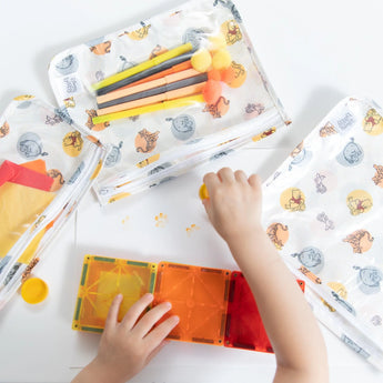 A child plays with colorful magnetic tiles near Bumkins Clear Travel Bag: Winnie and Friends and pom-pom topped markers.