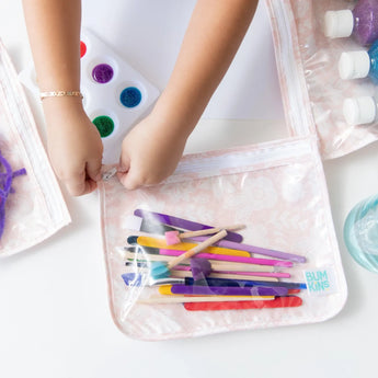 A child zips a Bumkins Clear Travel Bag 3-Pack: Lace with art supplies, surrounded by paint pots and glitter jars. TSA-compliant.