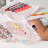 A child reaches for crayons beside Bumkins Clear Travel Bag 3-Pack: Lace, filled with vibrant colors on the table.