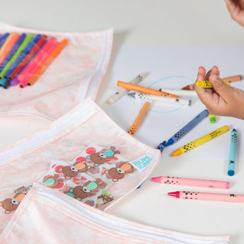 A child draws with colorful markers, surrounded by patterned pencil cases and Bumkins Lace Clear Travel Bag 3-Pack.