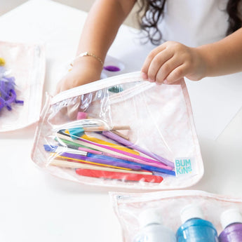 Child placing colorful pens in TSA-compliant Bumkins Clear Travel Bag 3-Pack: Lace on white table with art supplies.