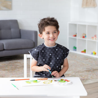 A child paints at a table in a Bumkins Mickey Mouse Icon B+W smock, with a cozy couch in the background.