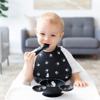 In their high chair, the toddler smiles in a Bumkins Silicone Bib: Mickey Mouse Faces, holding a spoon with a bowl in front.