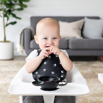 A baby in a high chair wears a Bumkins silicone bib with Mickey Mouse faces, enjoying snacks from a black bowl. A grey sofa and plant sit behind.